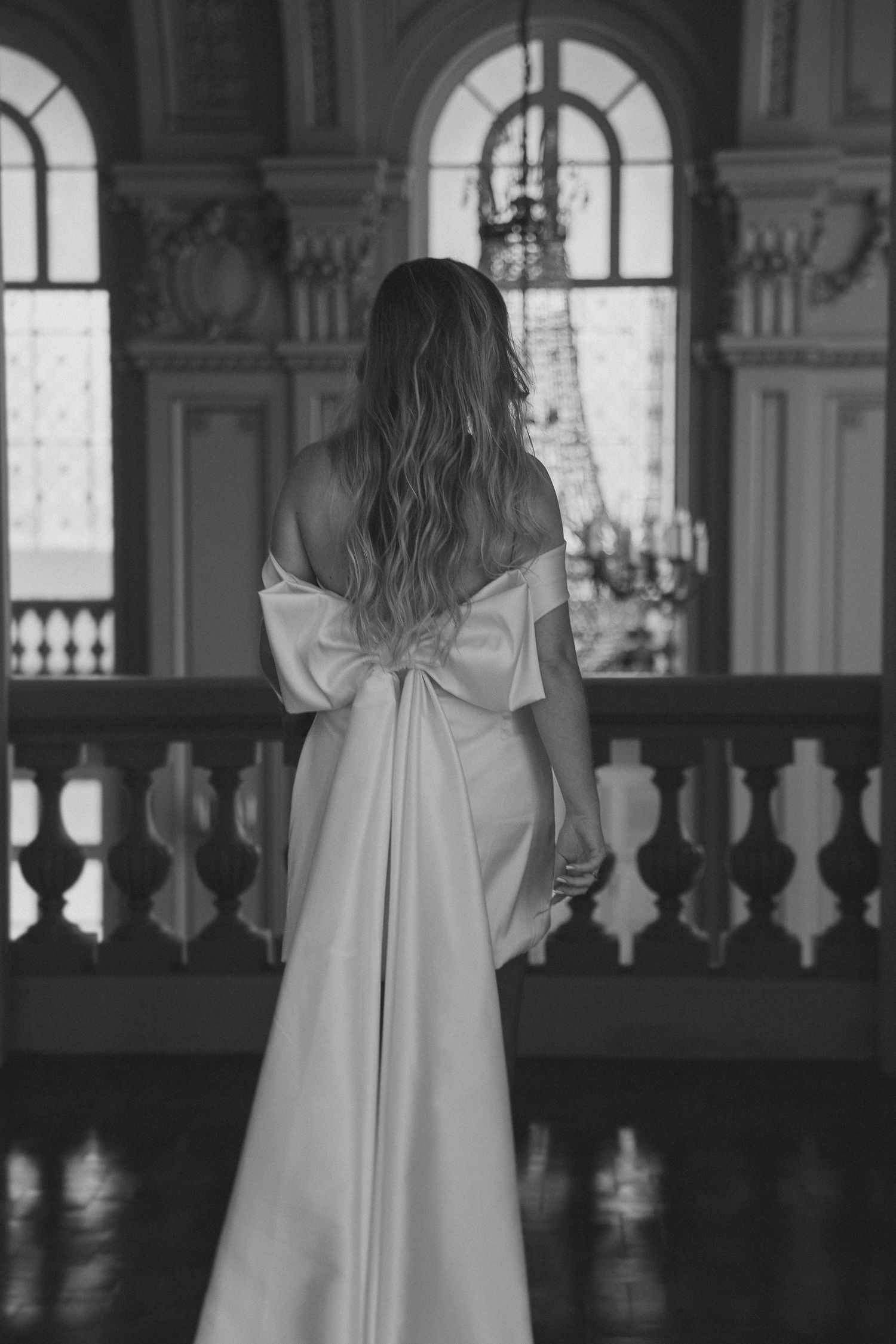 A bride in a white wedding dress with a large bow detail stands at an elegant balcony with arched windows.