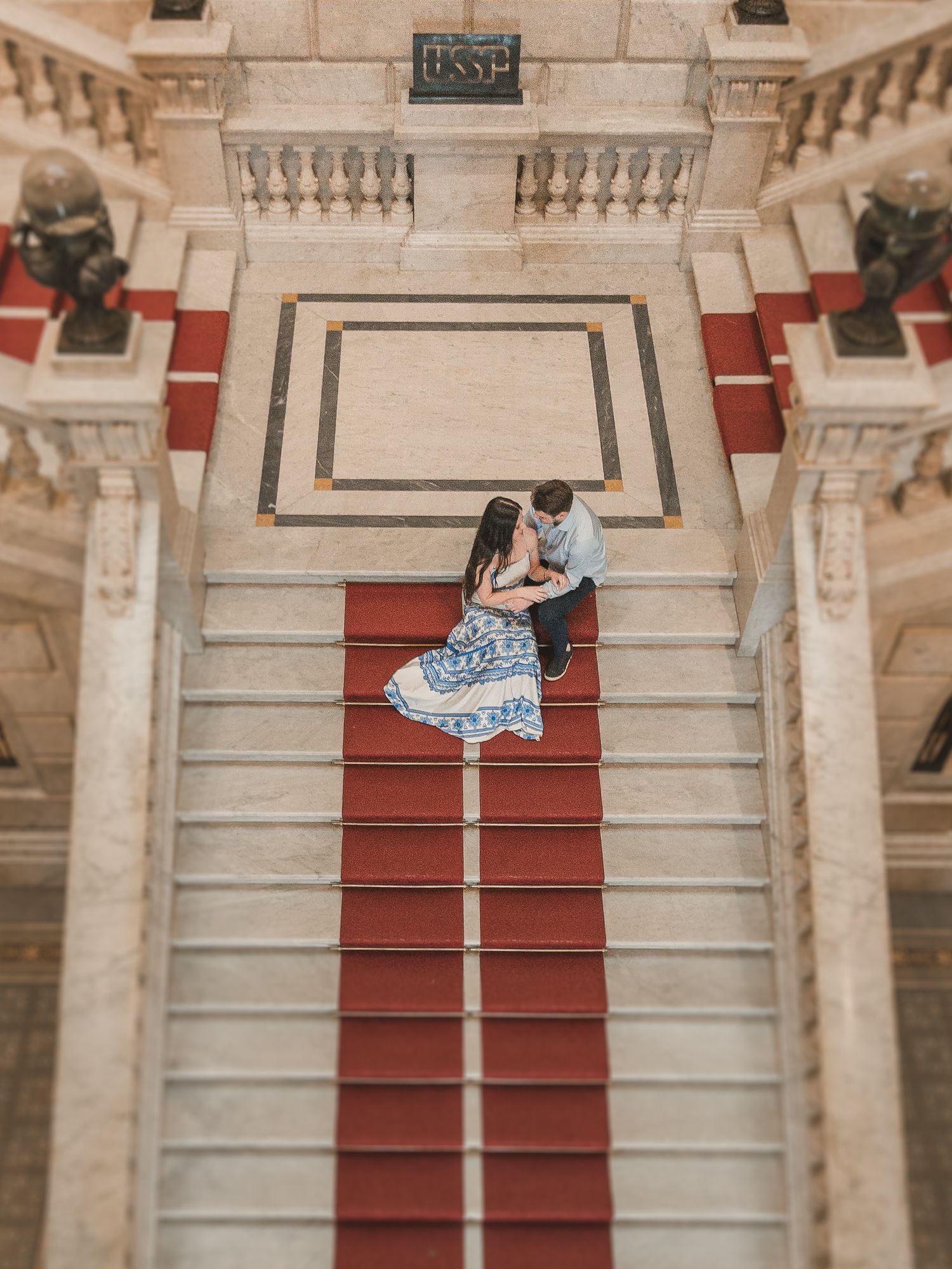 A romantic couple stands on a grand staircase with red carpet and ornate stone balustrades in an elegant interior setting.