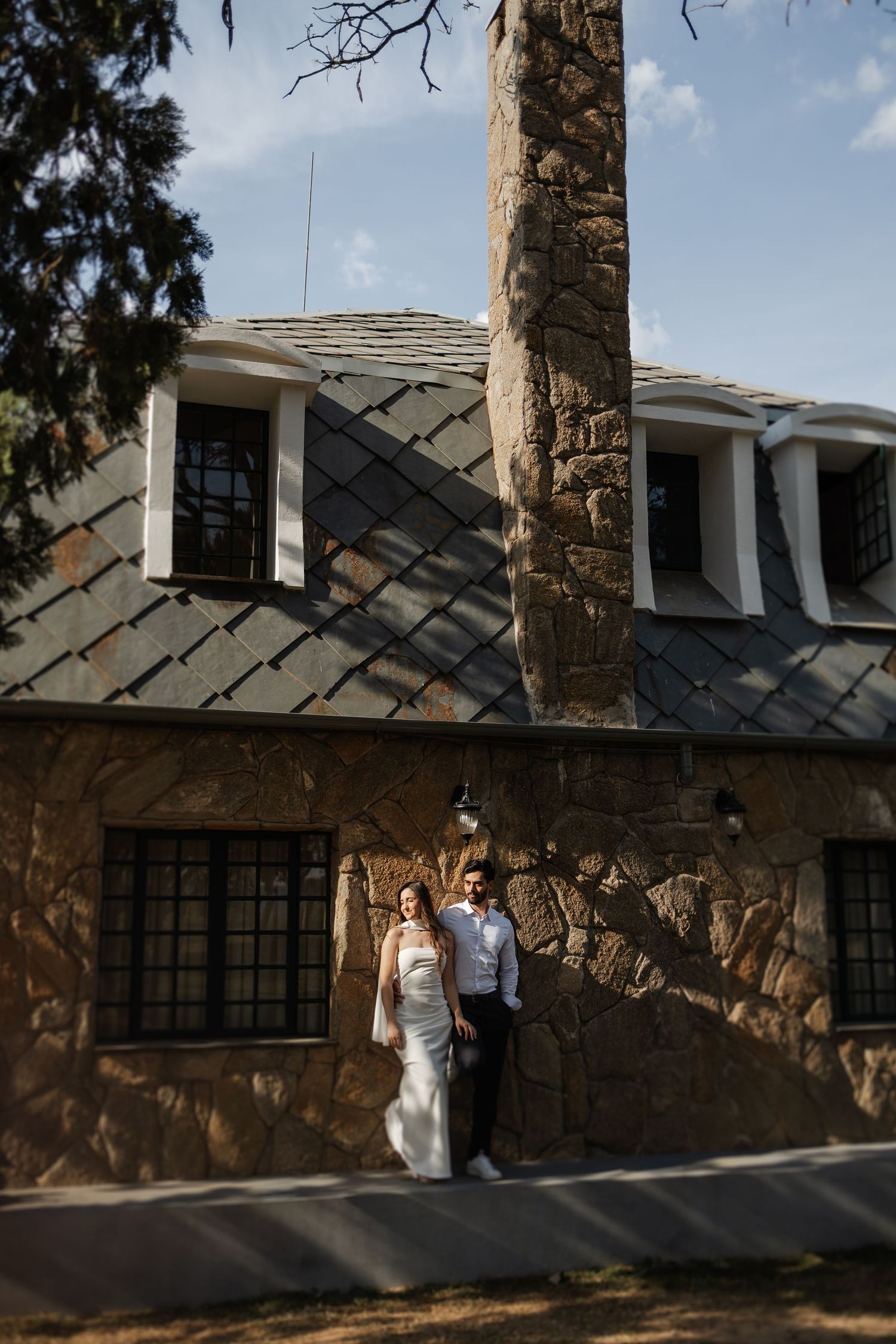 A couple stands in front of a rustic stone house with a grey slate roof and dormer windows on a sunny day.