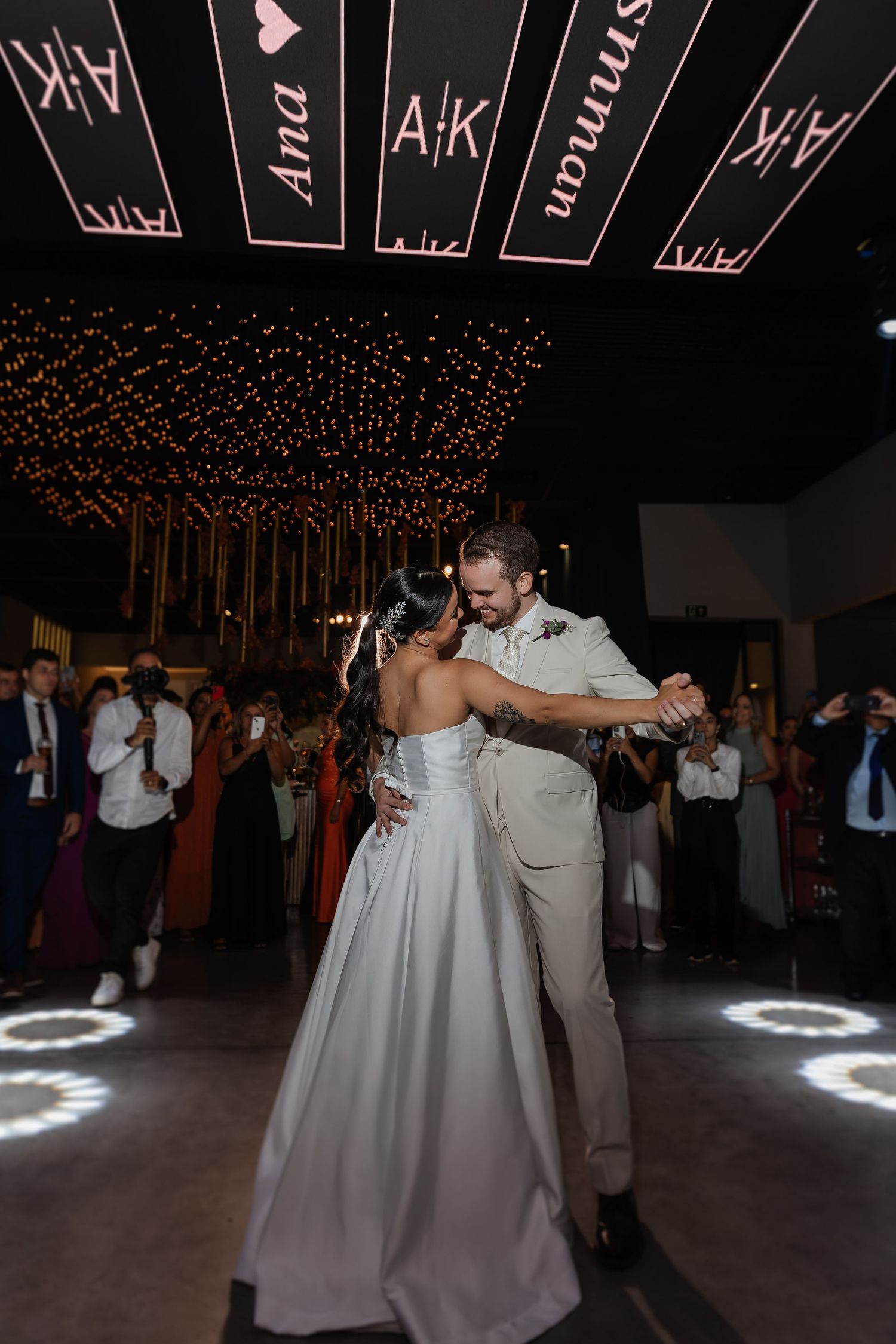 A couple shares their first dance at a wedding reception under strings of twinkling lights and modern black ceiling decor.