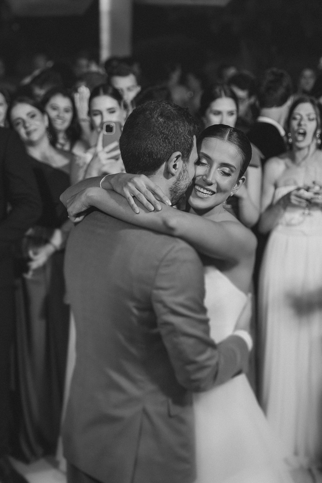A joyful first dance moment at a wedding reception captured in black and white with bridesmaids watching in background.