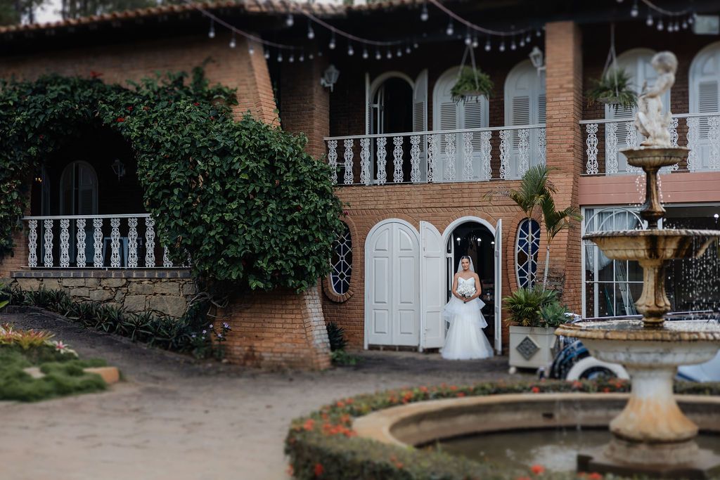 Bride standing in elegant white dress outside Mediterranean-style venue with fountain and ivy-covered walls.