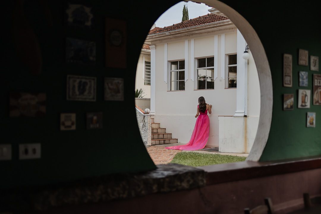 Through a circular archway, a figure in a flowing pink dress stands on a white porch of a colonial-style building.