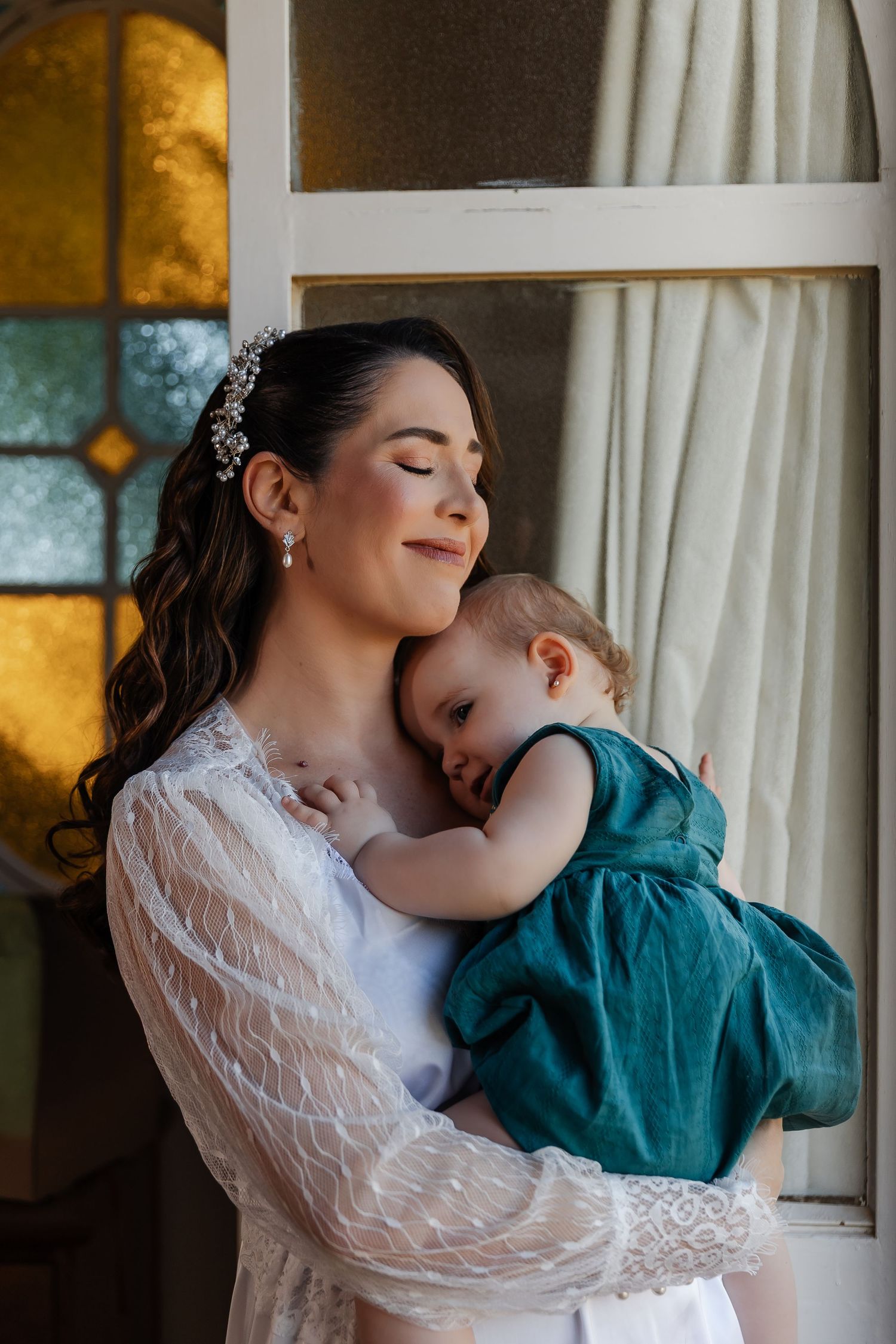 A tender moment between parent and child in white lace and teal clothing by a window with soft lighting.