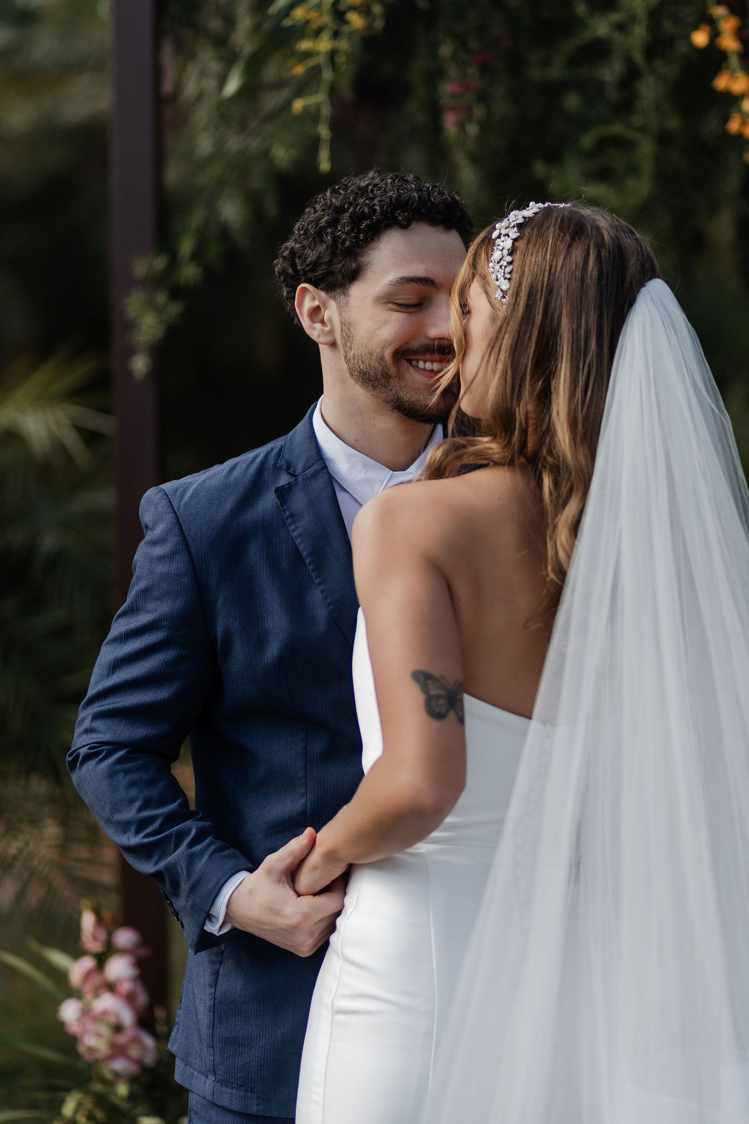 Romantic wedding couple embraces tenderly during outdoor garden ceremony with flowing white veil and navy suit.