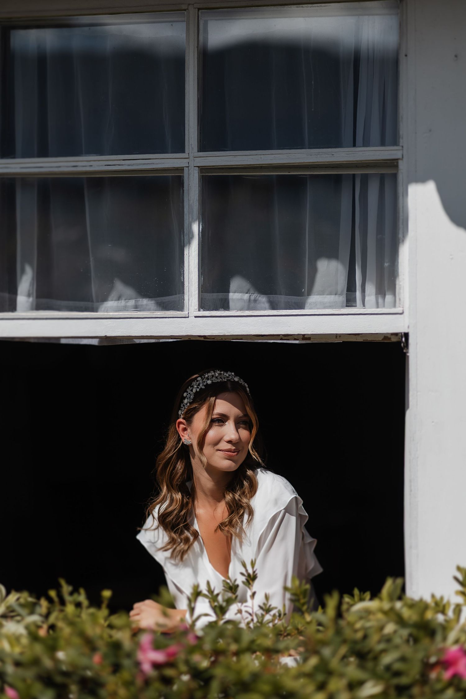 Woman wearing a white shirt poses by window with garden flowers visible in foreground on a sunny day.