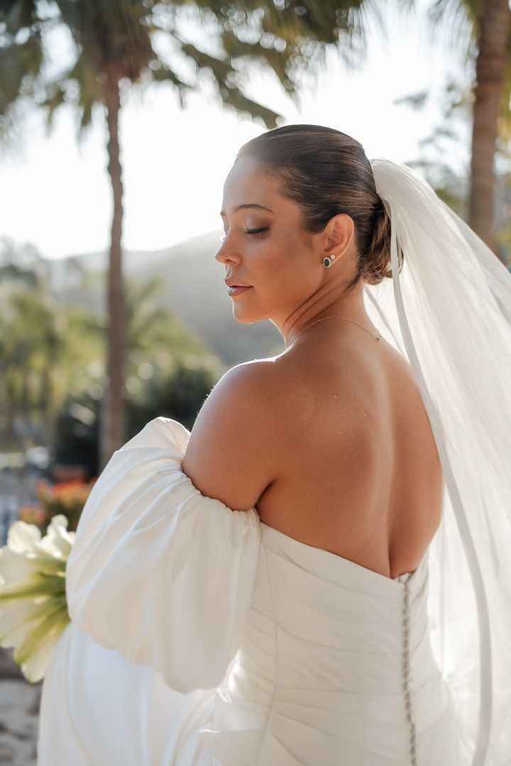 Elegant bride in off-shoulder wedding gown and flowing veil poses in tropical garden setting.