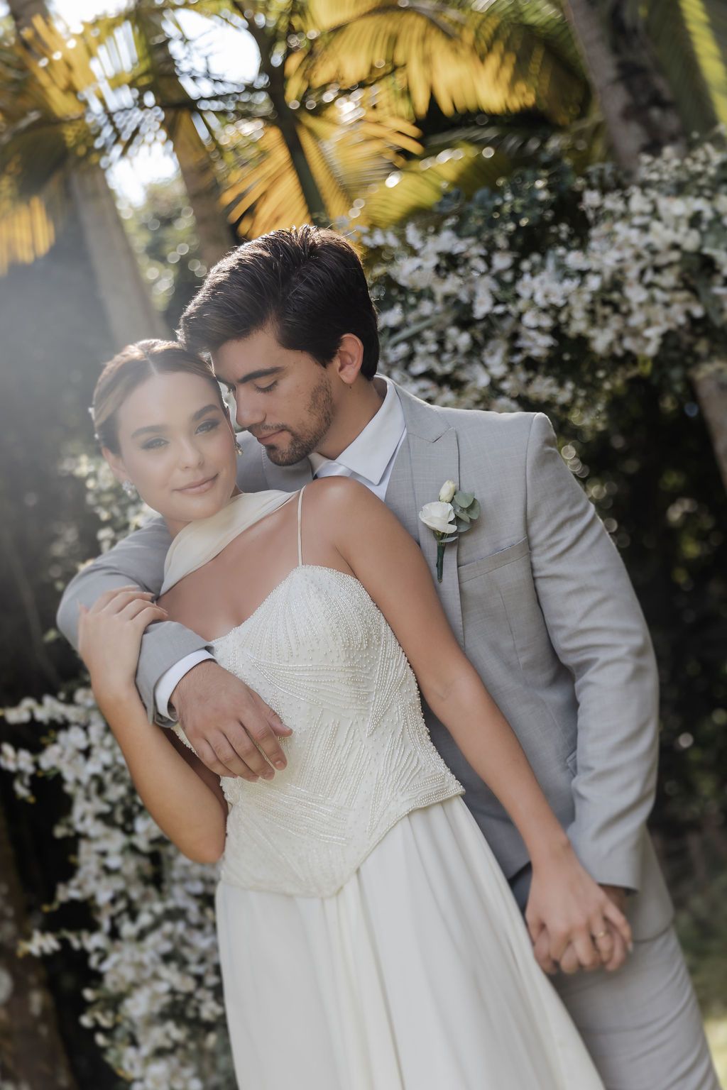 A romantic wedding photo of newlyweds embracing outdoors with sunlight filtering through trees and white flowers in background.