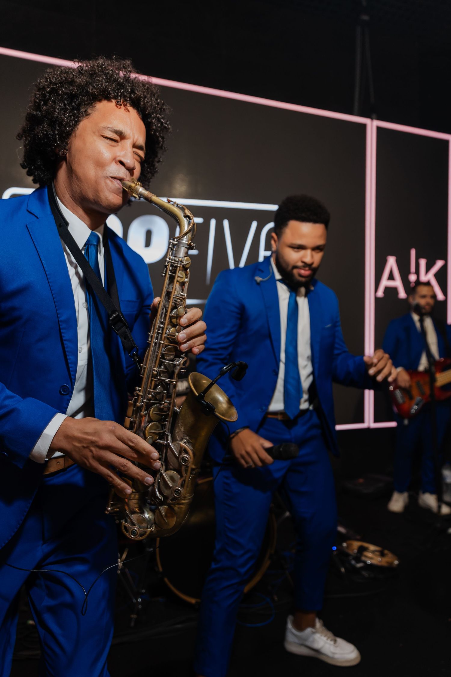 Musicians in matching royal blue suits perform on stage with a saxophone player in the spotlight.