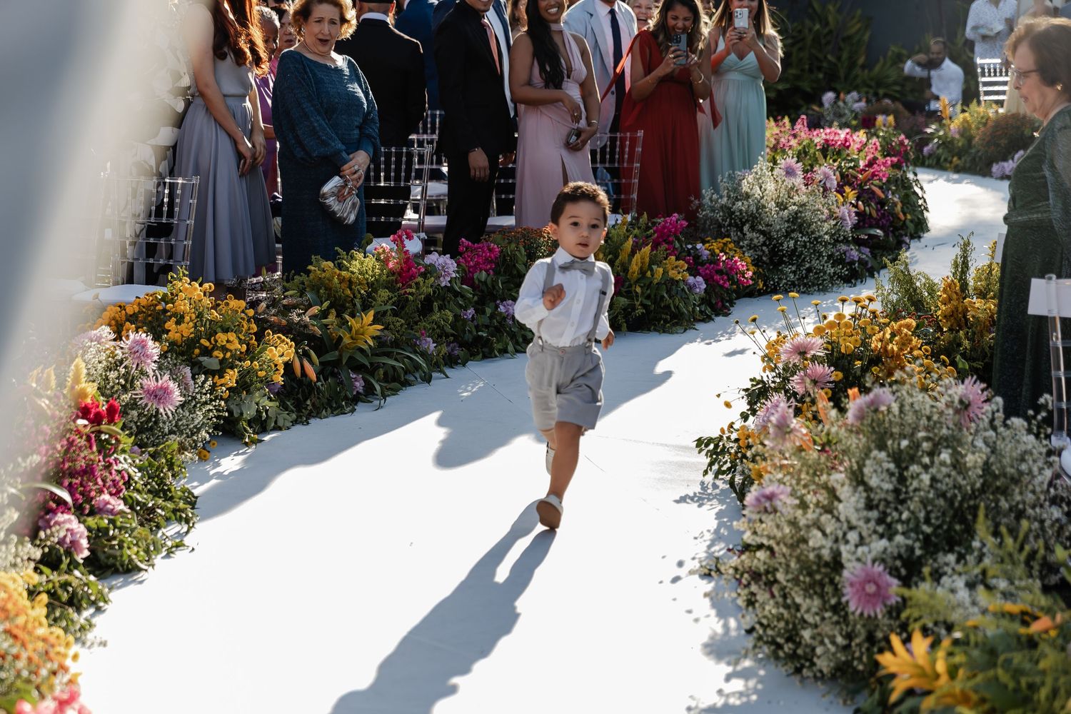 Young child walks down a white aisle lined with colorful flowers while guests look on during an outdoor ceremony.