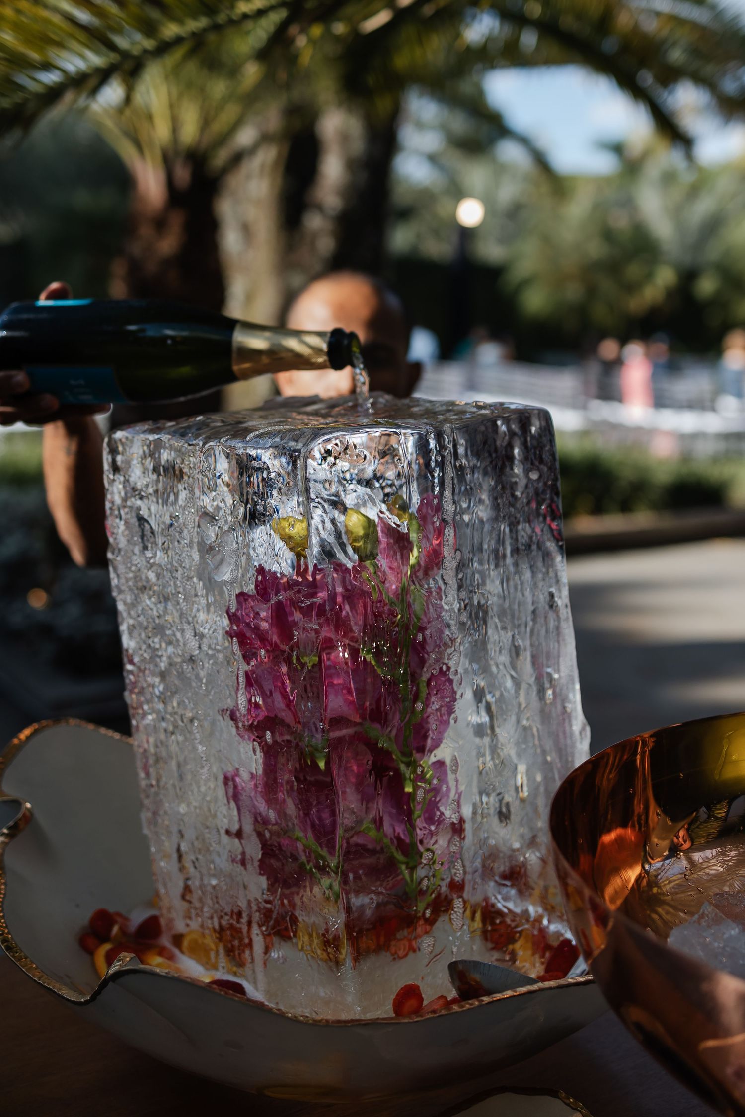 A beautiful pink flower frozen inside a large crystal-clear ice block illuminated in an outdoor setting with palm trees.