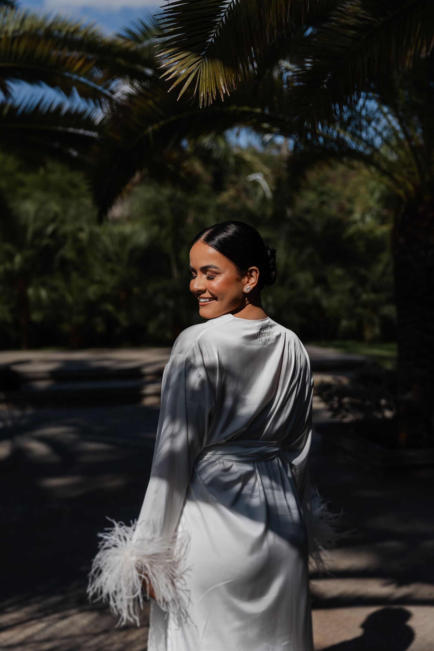 casamento em Belo Horizonte, casamento ao arA figure in a white silk robe with feathered cuffs stands under palm trees on a sunny afternoon.