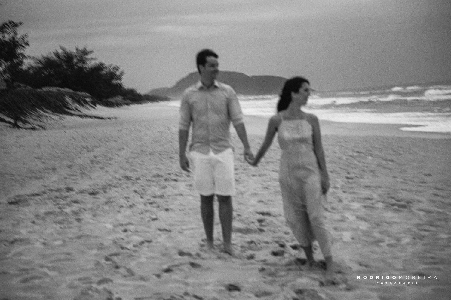 A couple walks hand in hand along a serene beach in black and white photography.