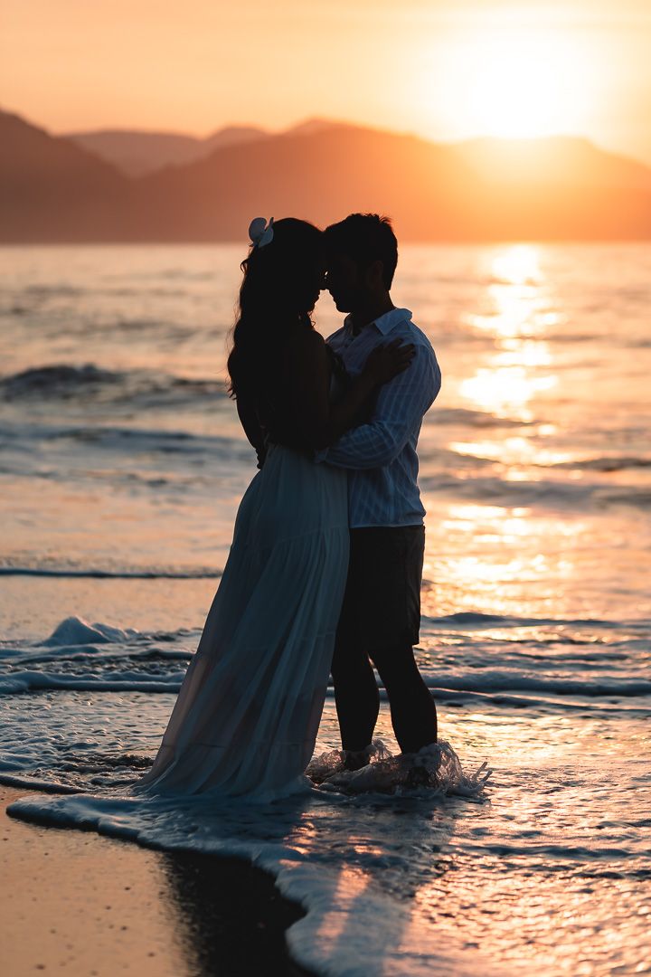 Silhouetted couple embracing on beach at sunset with waves lapping at their feet and golden light in the background.