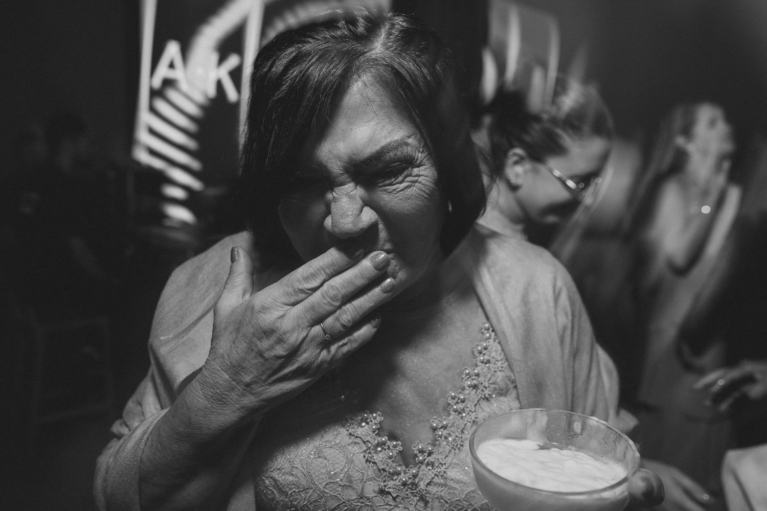 A black and white photograph shows someone laughing with hand over mouth while holding a cup at a social gathering.