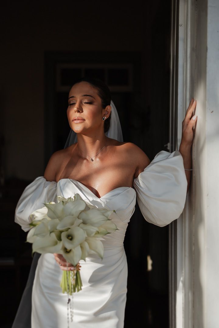 A bride in an elegant white strapless gown with puff sleeves holds a bouquet of calla lilies while standing by a window.