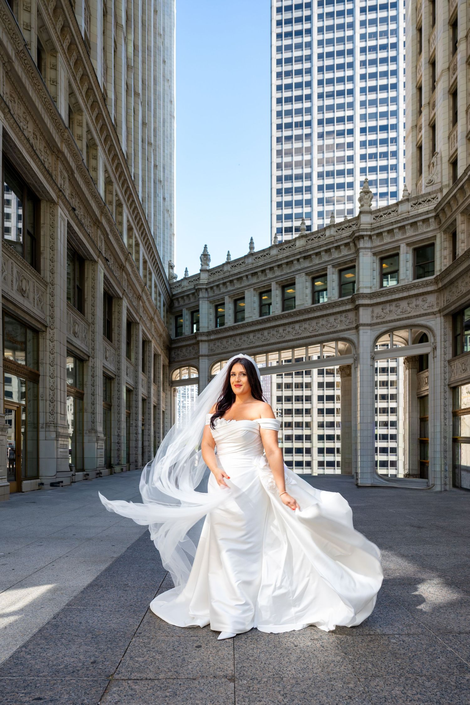 A bride in a flowing white gown poses dramatically in an elegant architectural setting with tall columns and buildings.