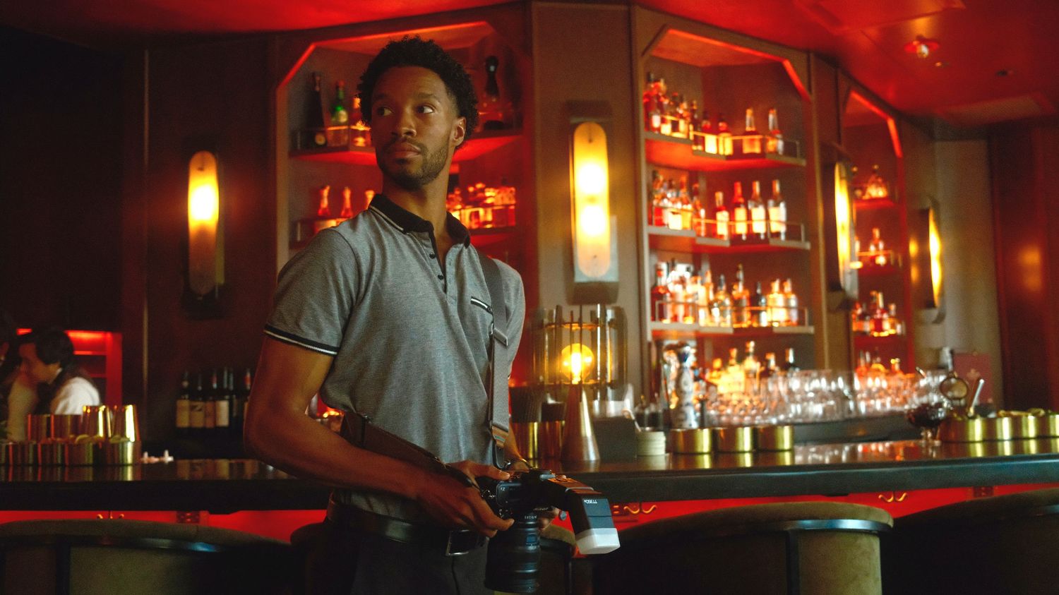 Red-lit bar interior with bottles and glasses displayed on shelves, creating a moody nightclub atmosphere.