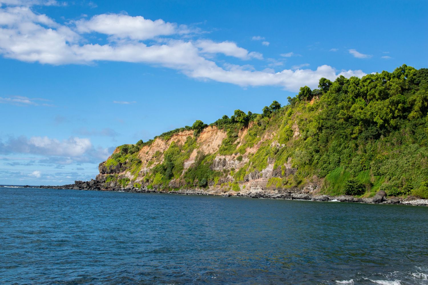 Dramatic coastal cliffs with lush green vegetation rise above blue waters under a bright sky.