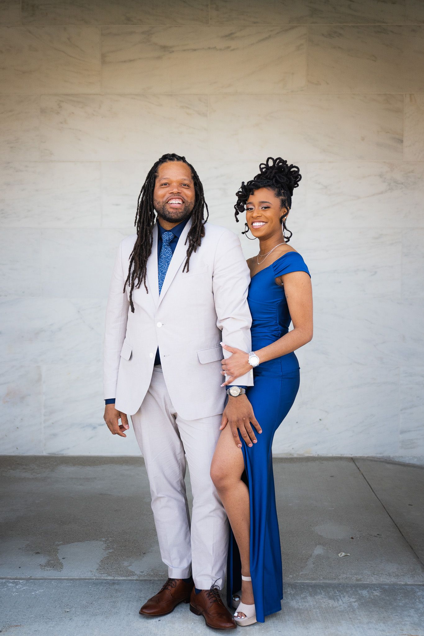 A couple in elegant blue and white formal attire poses against a white wall.