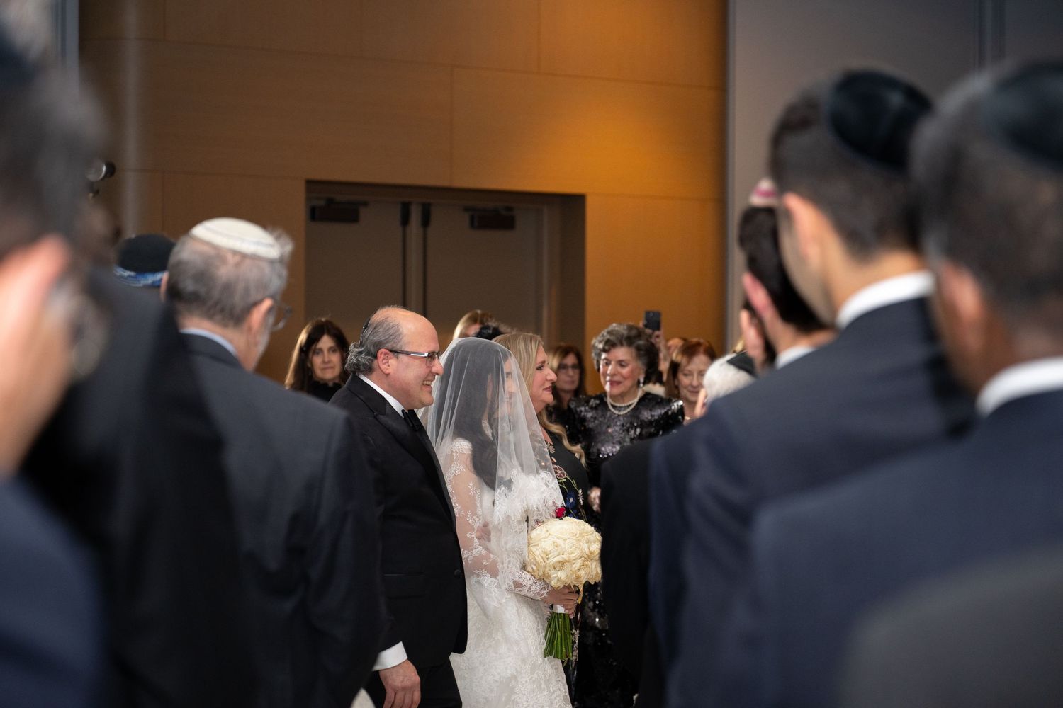 A bride and groom exchange rings during an indoor wedding ceremony surrounded by guests in formal attire.