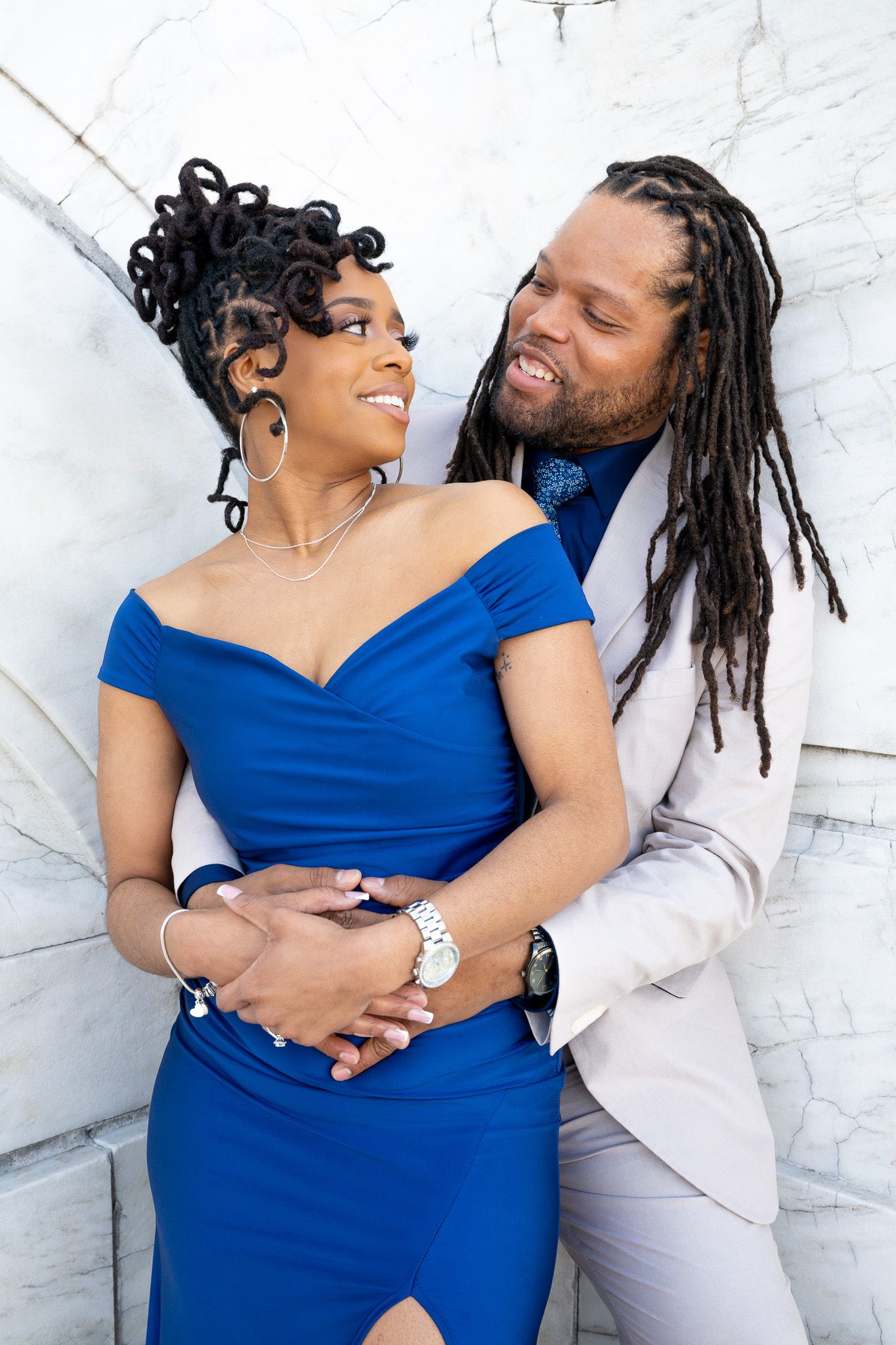 A couple in blue dress and light suit embrace against a white wall backdrop.