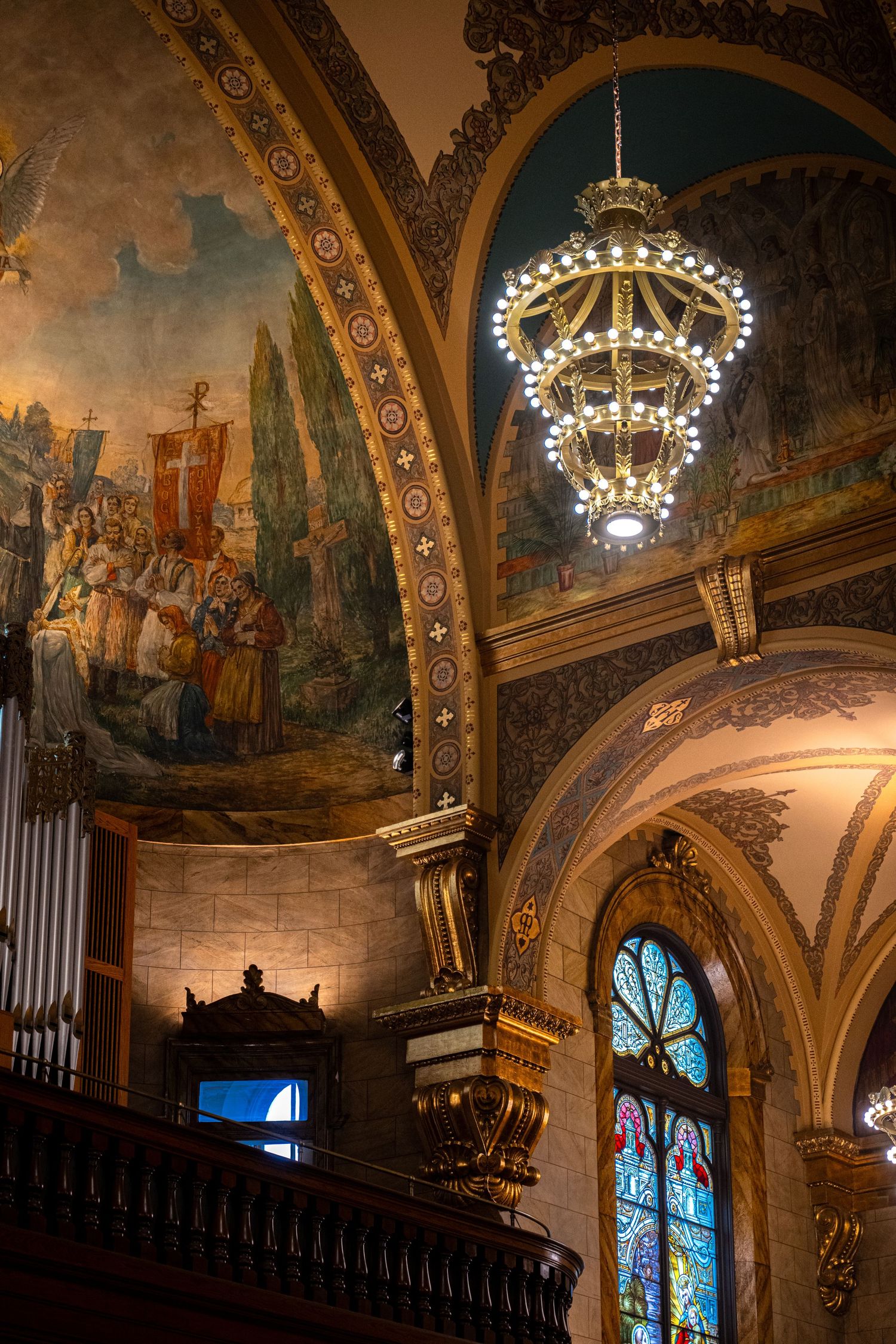 Ornate cathedral interior with vaulted ceilings, chandeliers, and painted murals depicting historical scenes in golden light.