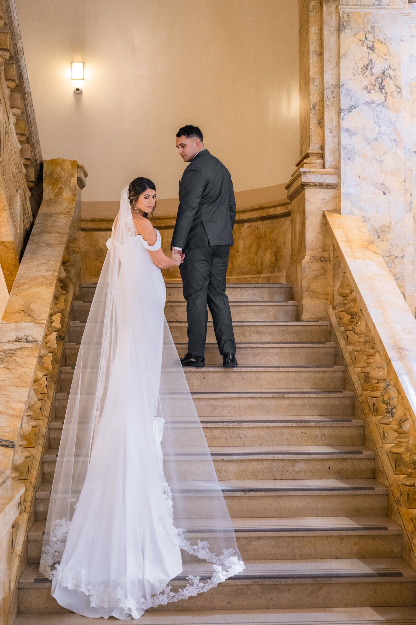 A couple in wedding attire climbs an elegant stone staircase with ornate golden railings.
