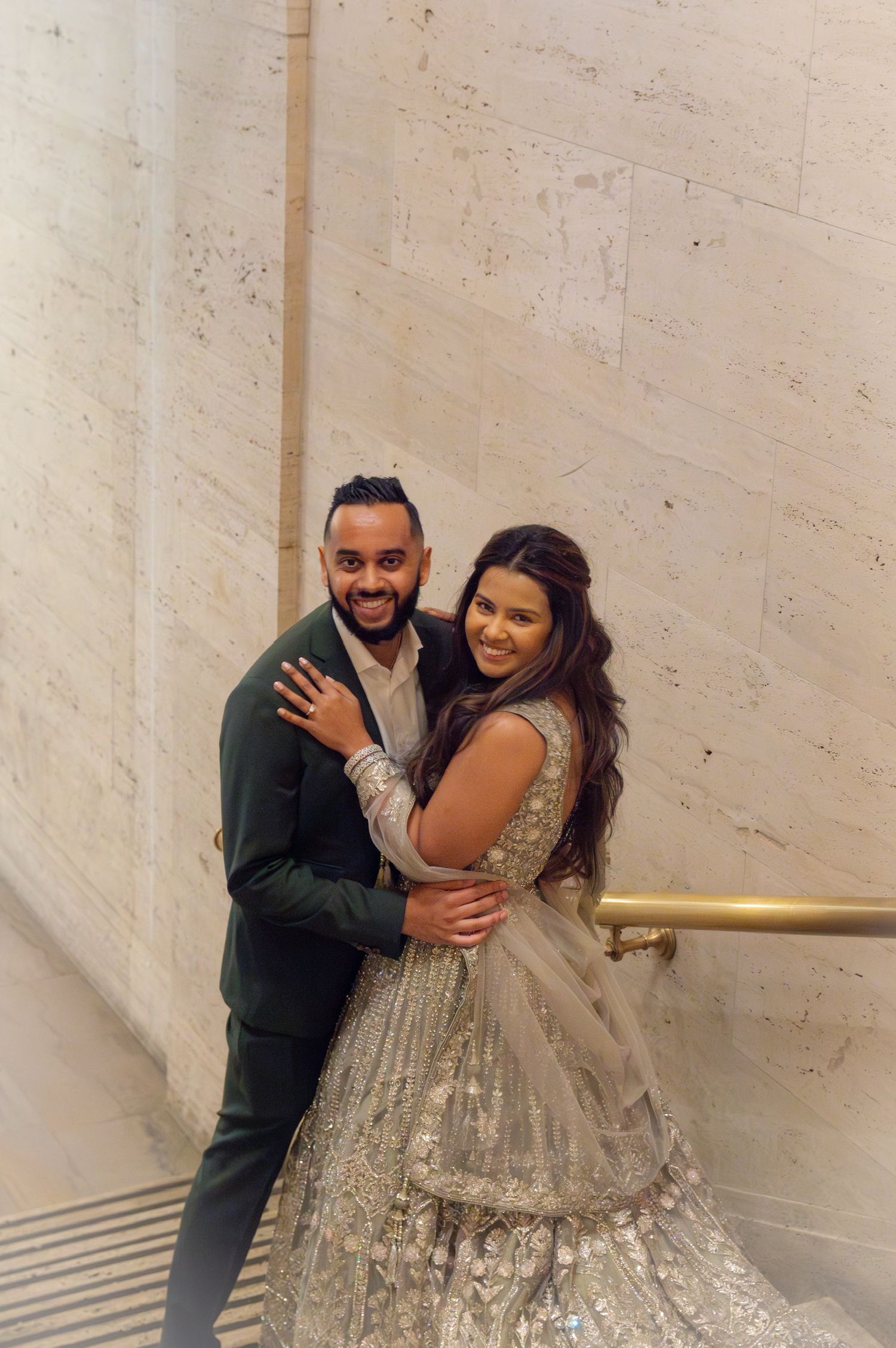 Couple in formal attire embracing against light stone wall with metallic handrail.