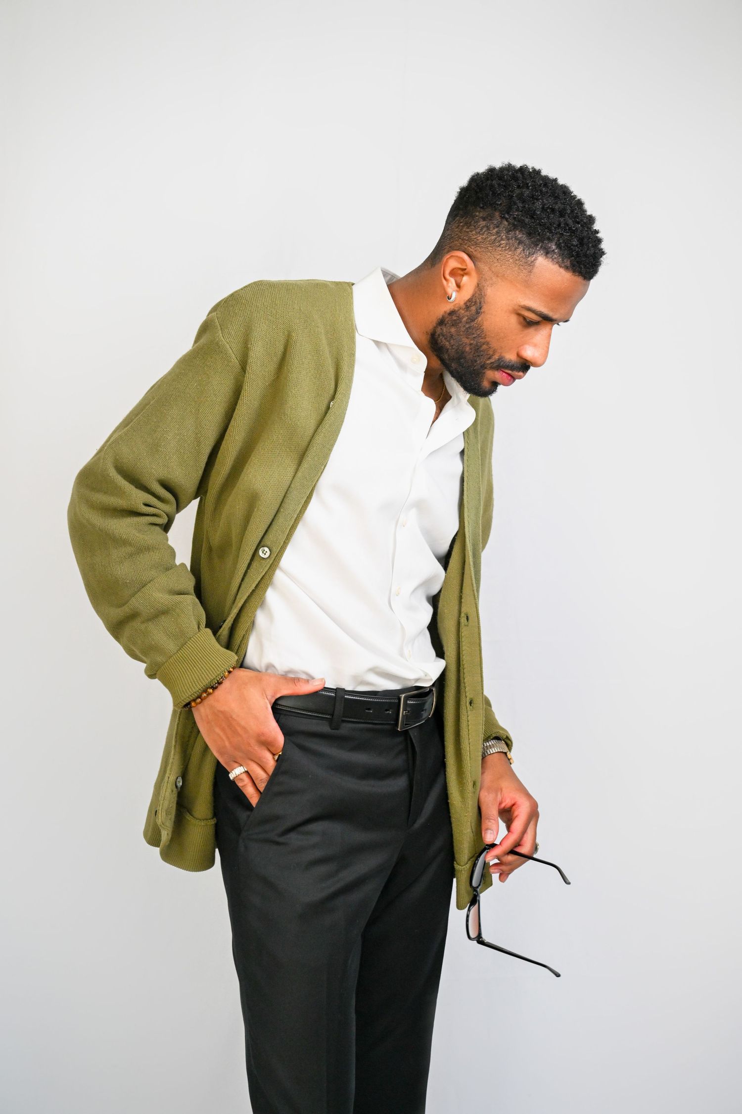 A person wearing a green cardigan and white shirt adjusts their belt while standing against a white background.