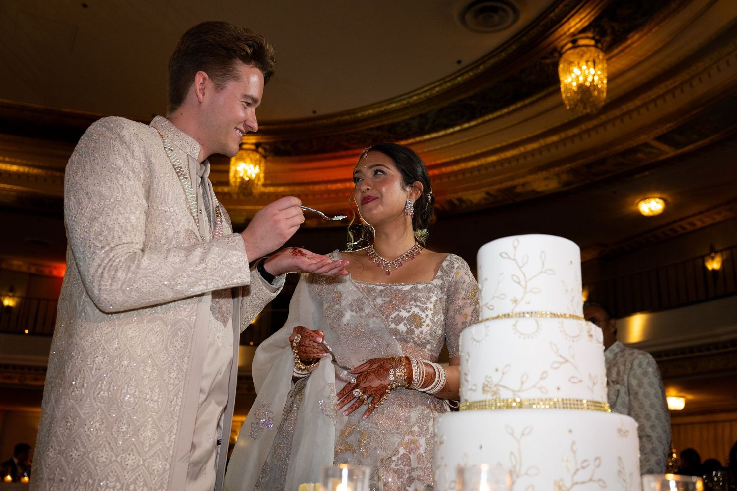 A bride and groom share a slice of wedding cake at their elegant reception with ornate lighting and decor.