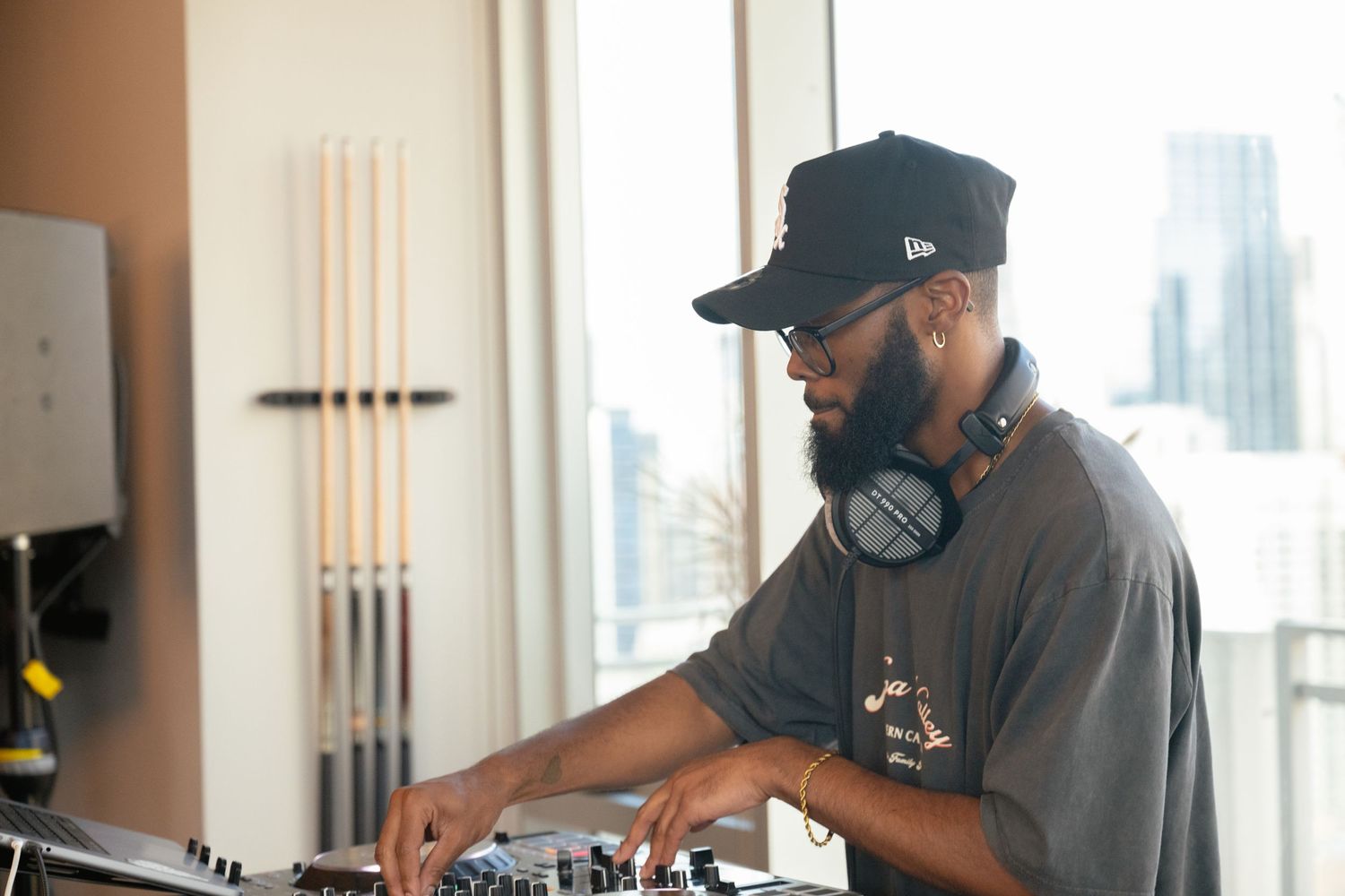 DJ performing at turntables near window with city skyline view.