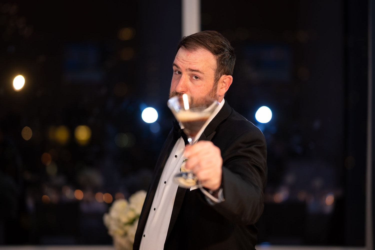 A person in a black suit holds a lit cigarette at night with blurred city lights in the background.
