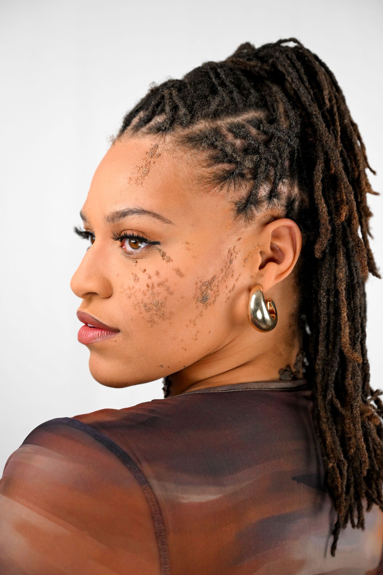 Side profile showing dreadlocks hairstyle and peace symbol earring against white background.