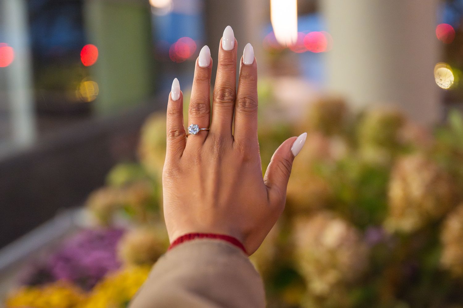 Close-up of an elegantly manicured hand with white nails and a delicate ring against a blurred floral background.