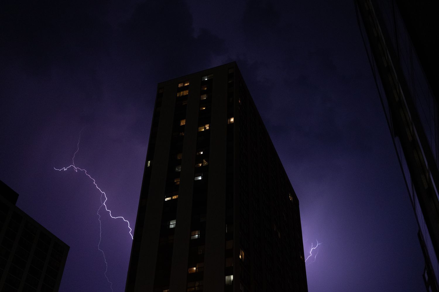 Lightning strikes illuminate the night sky behind a tall apartment building during a storm.