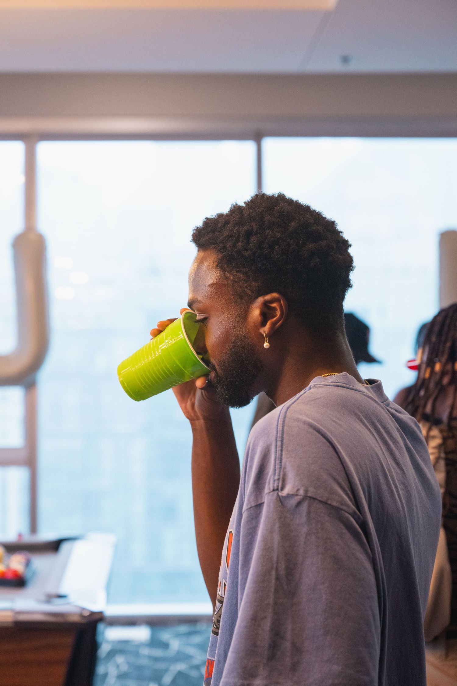 Person in gray shirt drinking from bright green cup against window backdrop.