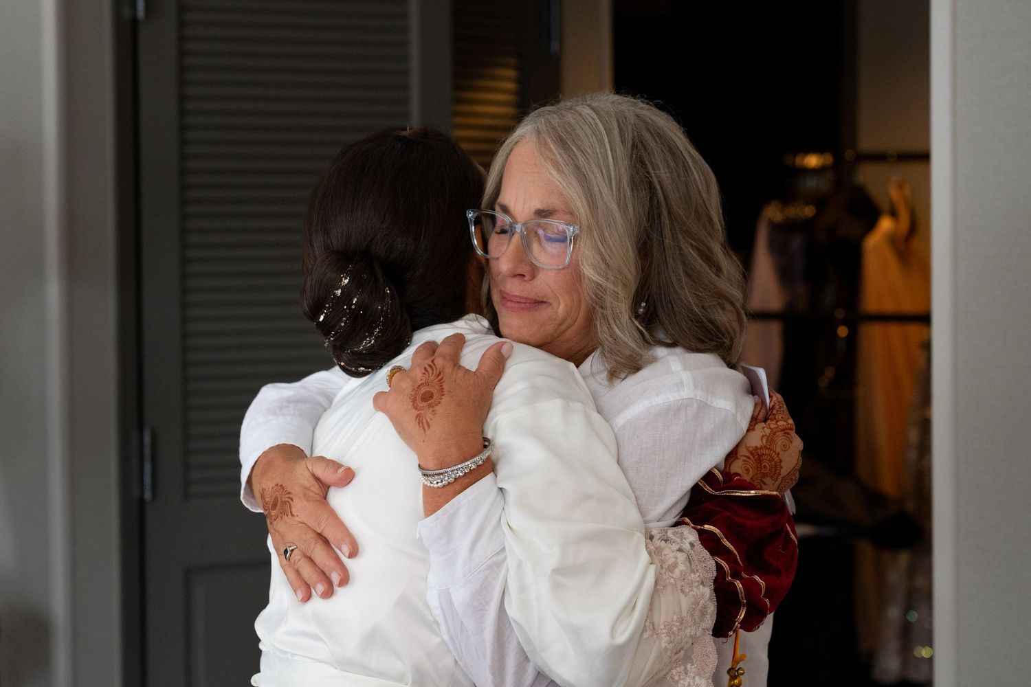 Two people share a tender embrace while wearing white clothing in a dimly lit indoor setting.
