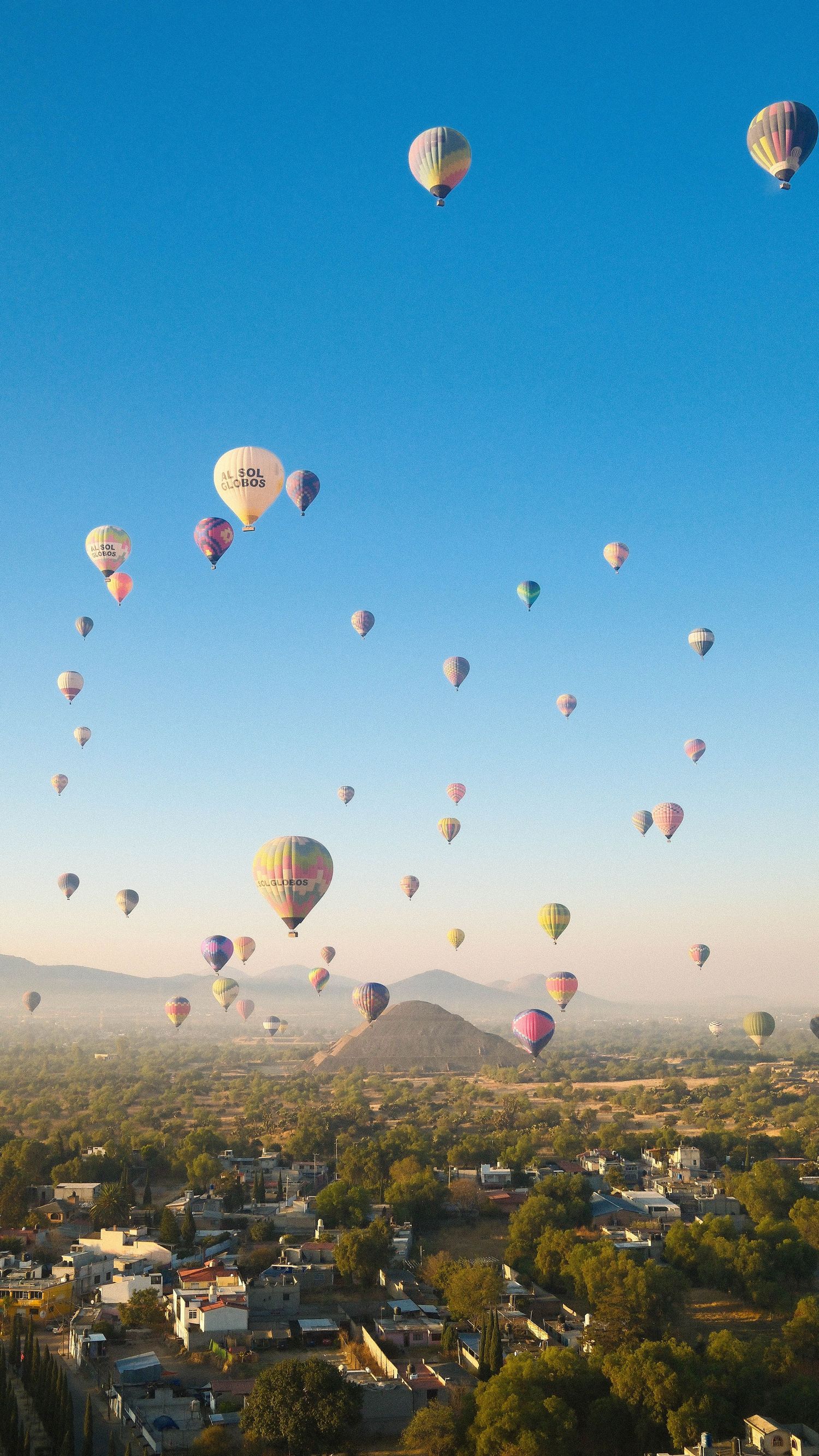 Multiple colorful hot air balloons float above a residential neighborhood against a bright blue sky and mountain backdrop.