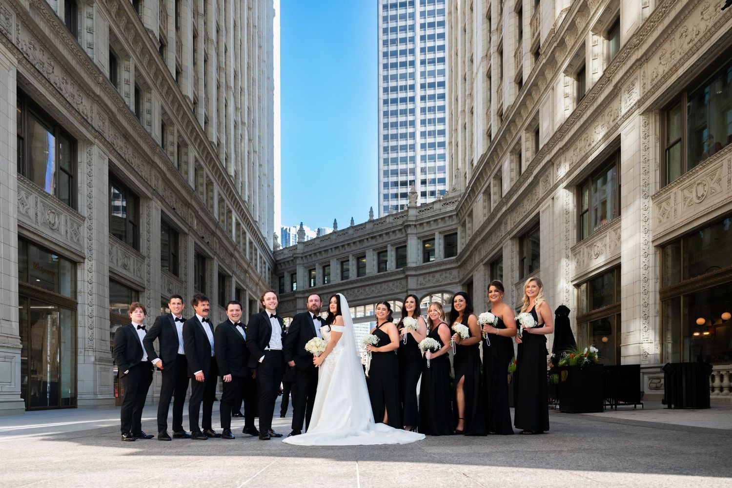 A wedding party poses between tall historic buildings in downtown Chicago on a sunny day.