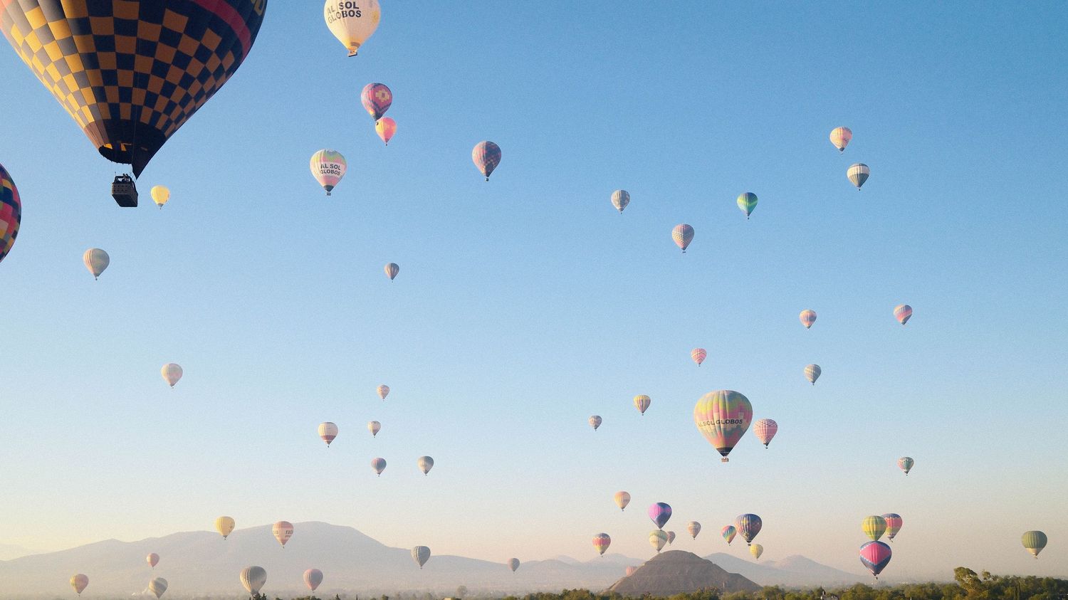Colorful hot air balloons float across a clear blue sky during a mass ascension at a balloon festival.