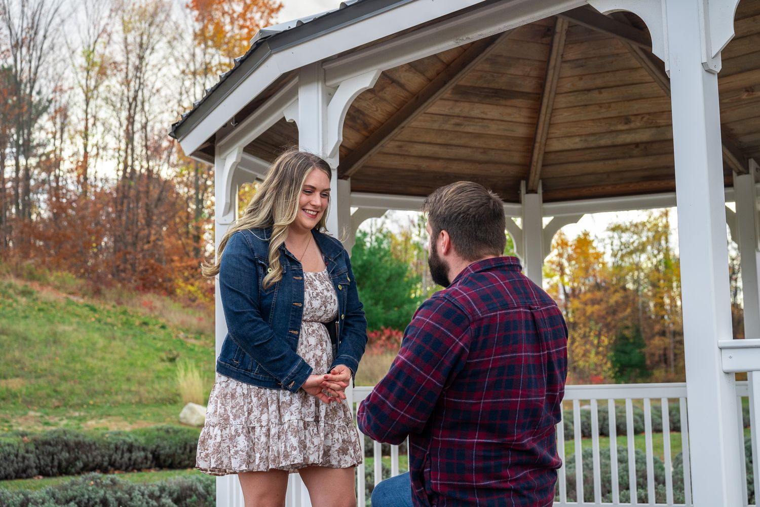 Proposal moment under wooden gazebo during autumn with colorful fall foliage in background.