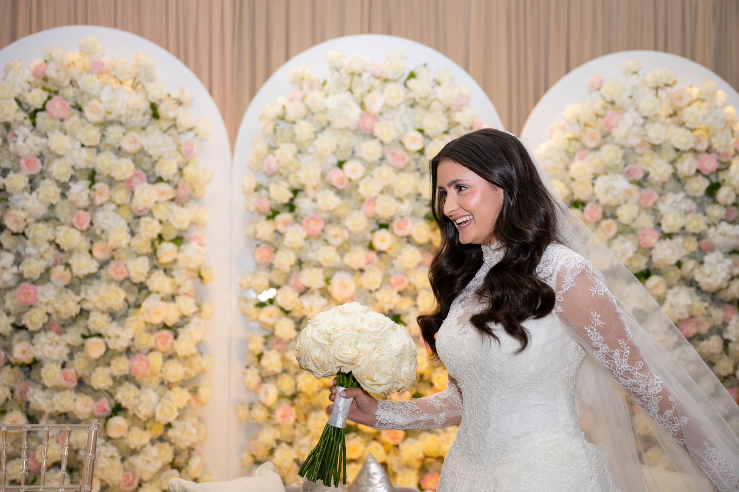 A bride in a white lace gown stands before an elegant floral wall arch display of cream and white roses.