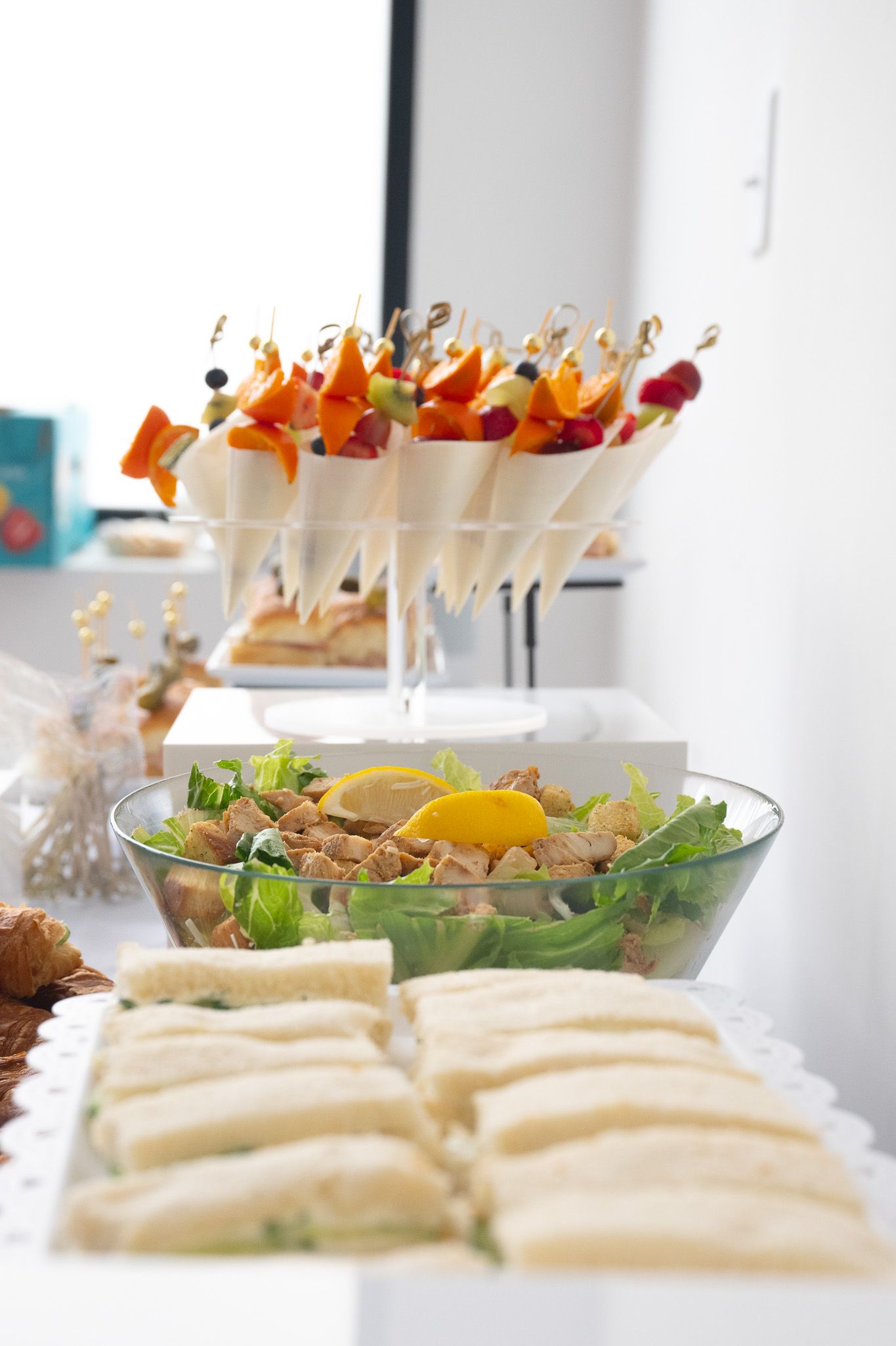 Catering spread featuring sandwiches, salad, and appetizer skewers on a white table.
