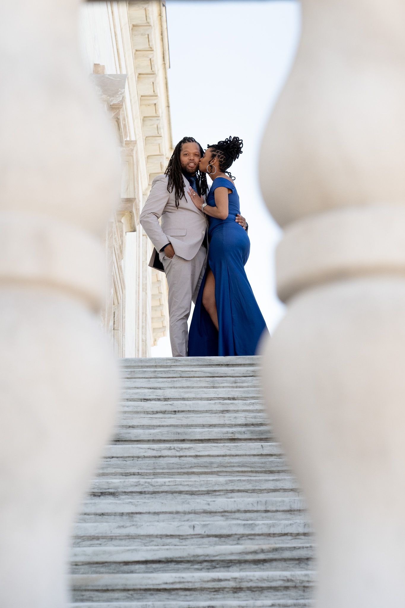 A couple in formal attire poses on white marble steps against a bright background.