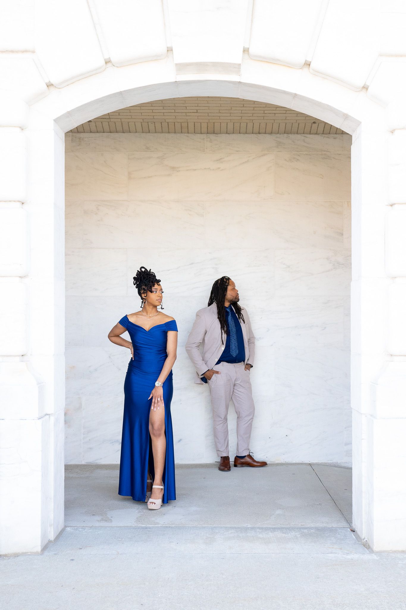 A couple poses playfully in blue and light gray formal attire against white archways.