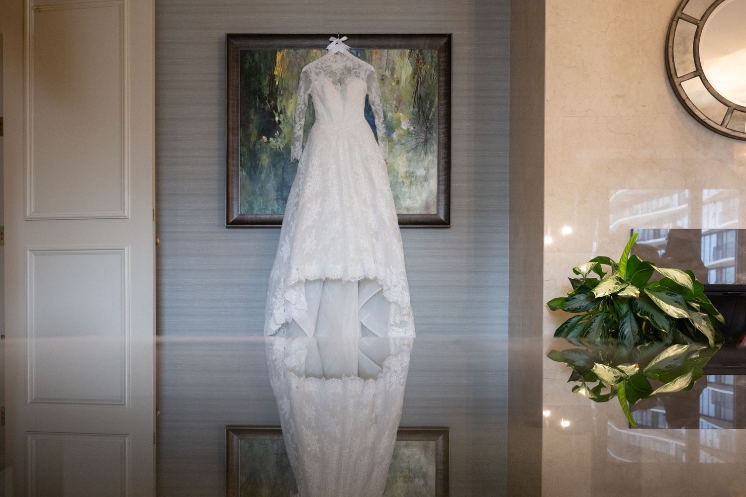 A white wedding dress with lace details hangs on display against a gray wall with potted plants and decor.