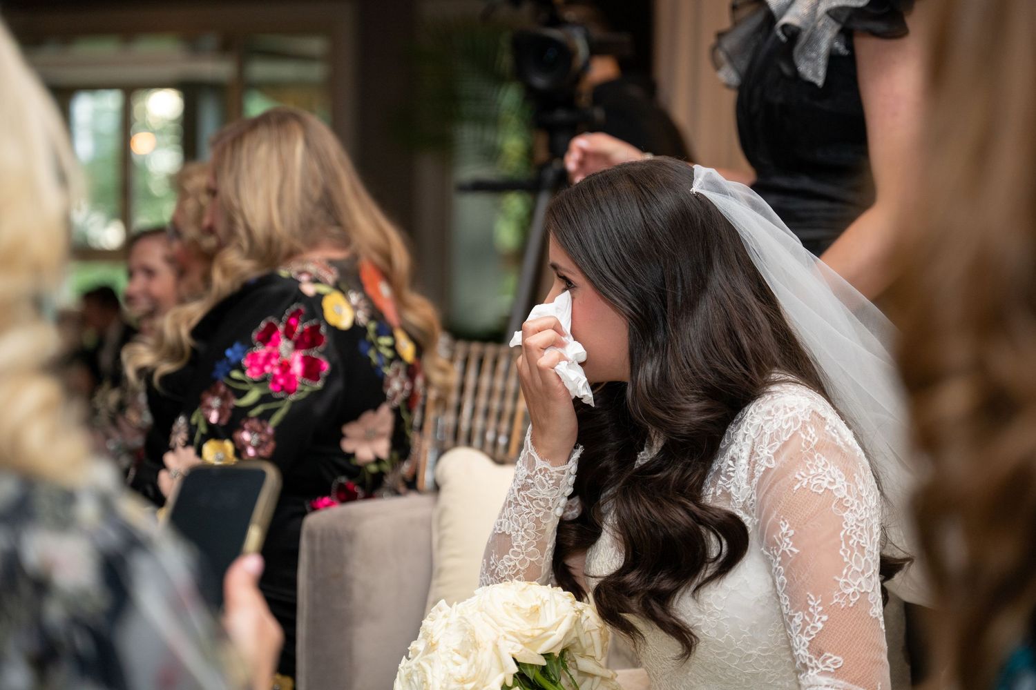 An emotional moment at a wedding as a bride in a lace gown wipes away tears while holding a white bouquet.
