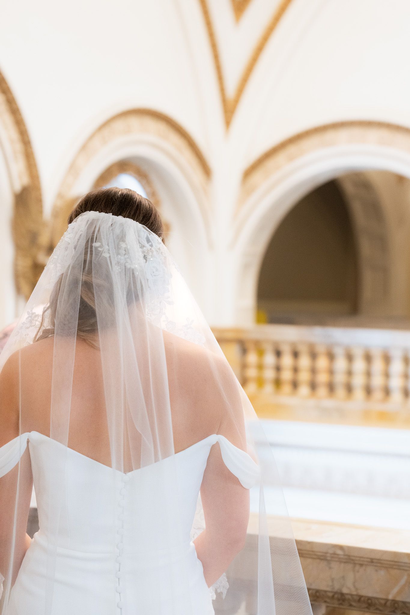 A bride in a white wedding dress and veil stands in an elegant arched hallway.