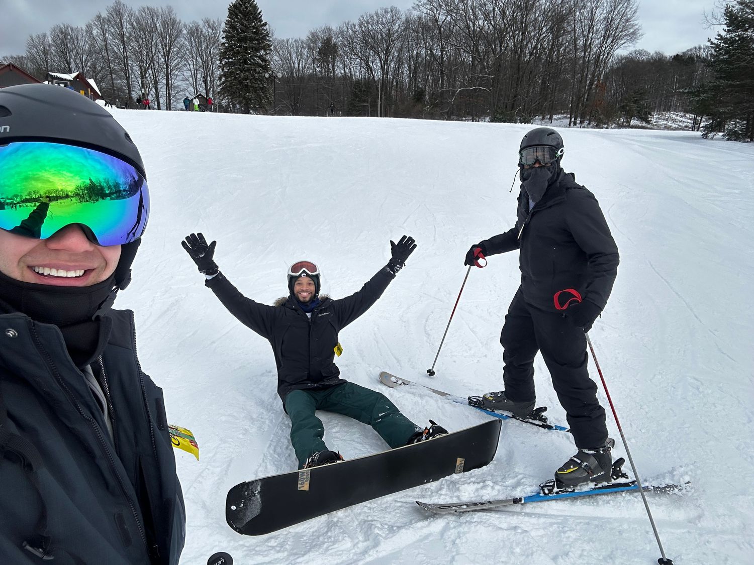 Three skiers taking selfie on snowy slope with one person lying down in background.